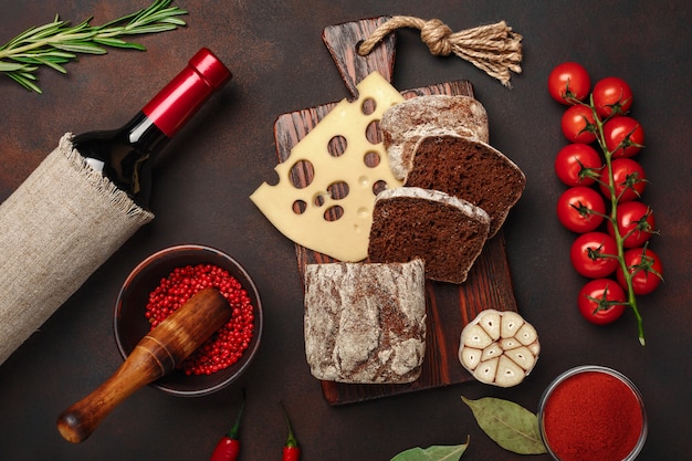 Maasdam cheese sliced on a cutting board with cherry tomatoes, black bread, garlic and rosemary, bottle of wine