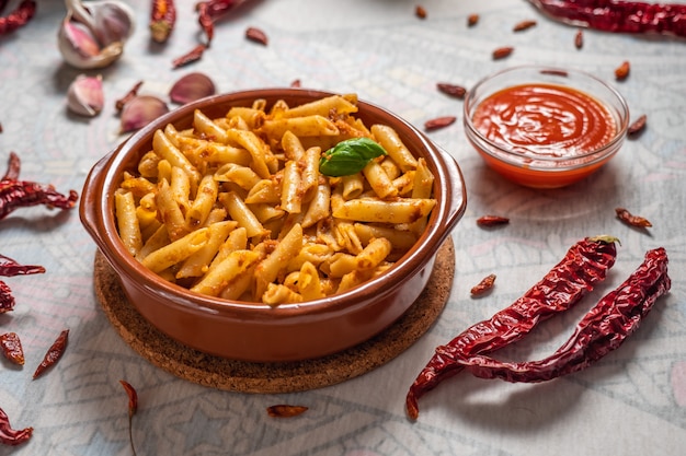 Macaroni with red pesto in a clay pan with some ingredients around on a white table.
