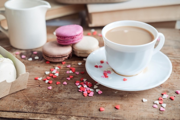 Macaroons and a cup of coffee a milk jug on a background of small hearts on a wooden background top view