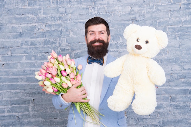 Macho getting ready romantic date. man wear blue tuxedo bow tie\
hold flowers bouquet. international womens day. surprise will melt\
her heart. romantic man with flowers and teddy bear. romantic\
gift.
