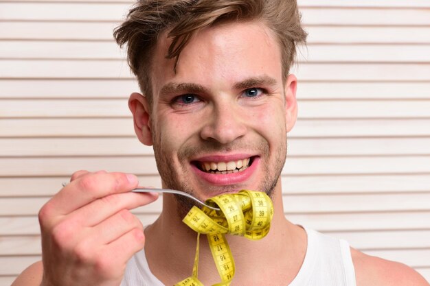 Macho met verleidelijk gezicht houdt zwarte kom met geel meetlint vast als voedsel. de mens beweert noedels op lichte houten plankenachtergrond te eten. man met sterke spieren in witte tanktop toont gekrulde tape.