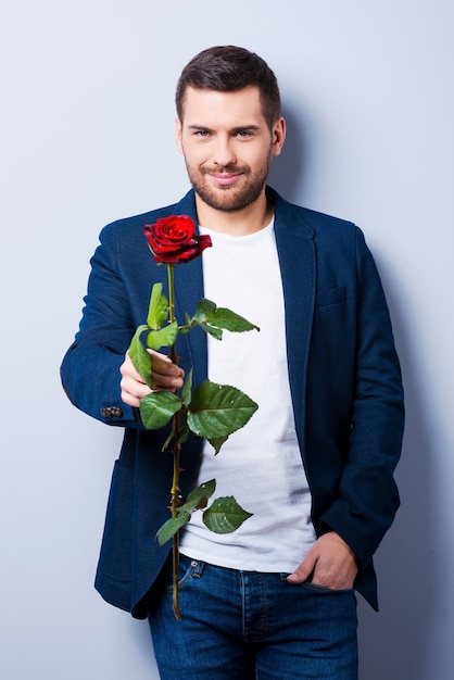 Macho and rose. Handsome young man giving a flower to you while standing against grey background