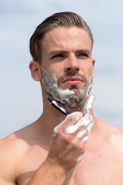 Macho scheren kin met scheermes man bedekt met scheerschuim schoonheid en gezondheidszorg concept mannelijk lichaam met sexy sterke torso Man met haren op heldere hemelachtergrond