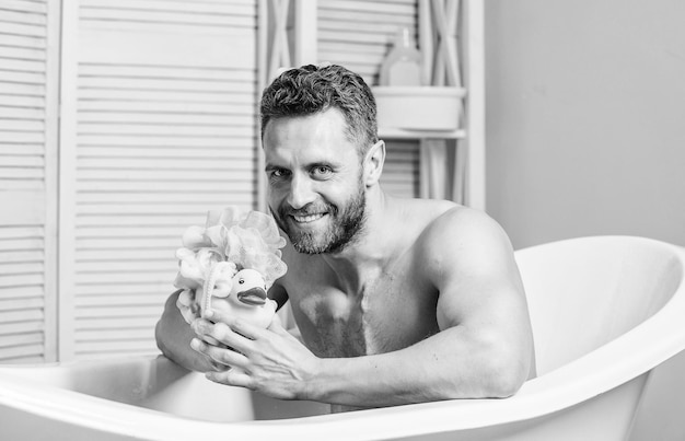 Macho with sponge take bath at home Taking bath with soap suds Beauty routine Relax and fun concept Daily bath helps beat depression Handsome muscular man relaxing bathtub with yellow duck