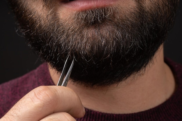Macro van een bebaarde man die pijnlijk grijs haar uit een dikke zwarte baard trekt met een pincet Mannenhaar en huidverzorging