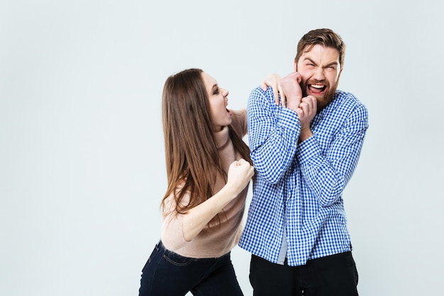 Mad aggressive young woman shouting and fighting with her husband over white wall