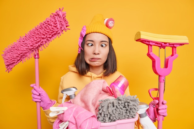 狂ったアジアの主婦は、家事をしている間、2つのモップを掃除に忙しくし、洗濯をしている間、愚かです。目は黄色のスタジオの背景の上に隔離された保護ゴム手袋を着用します
