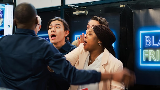 Mad diverse shoppers at store entrance going crazy for Black Friday promotions and deals, pushing through red tape. African american security agent managing crowd control during sales season.