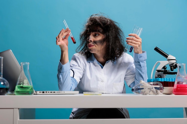 Mad scientist with goofy aspect in laboratory examinating liquid substance. Foolish wild looking crazy chemist analyzing glass test tubes filled with blood sample while sitting at desk in laboratory.