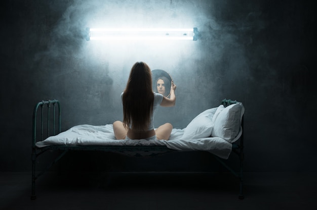 Mad woman with mirror sitting in bed, insomnia, dark room.. Psychedelic female person having problems every night, depression and stress, sadness, psychiatry hospital