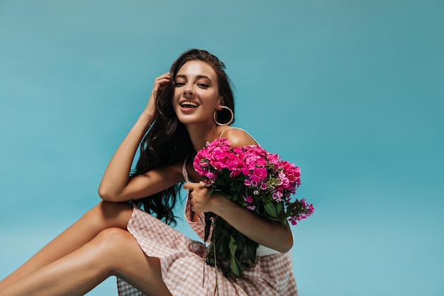 Magere gebruinde dame met lang krullend haar in modieuze roze outfit die lacht en een helder boeket op blauwe achtergrond vasthoudt