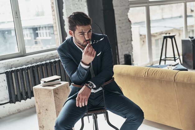 Magic look. Good looking young man in full suit putting on his eyewear while sitting on the stool