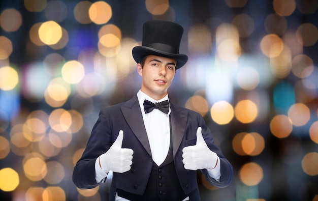 Photo magic, performance, gesture people and show concept - magician in top hat showing thumbs up over nigh lights background