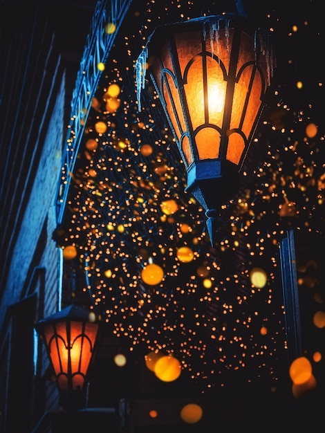 魔法のオールドストリートランタンが夜通りに輝いています。周りにたくさんの明るいライトがあります。家の壁にヴィンテージオールドストリートクラシックアイアンランタン。クリスマスやハロウィーンの魔法の妖精提灯。