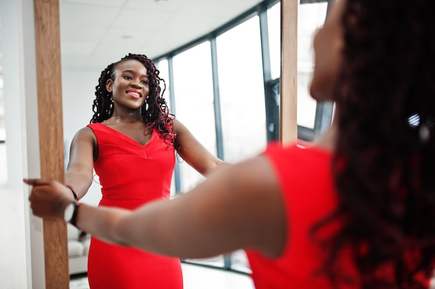 Magnifica giovane donna africana in lussuoso vestito rosso in un appartamento di lusso guarda allo specchio. bellezza, moda.