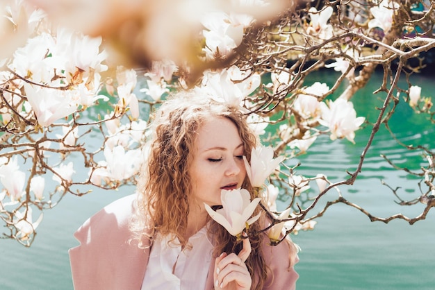 A magnificent young woman enjoys the spring weather in the park sniffs a blooming magnolia next to the pond