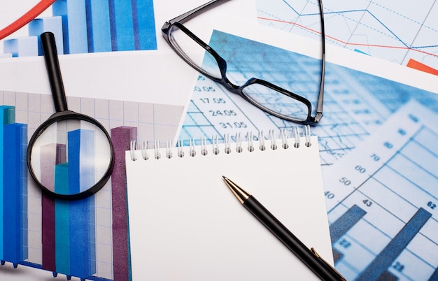 Magnifier, glasses, white notebook and pen on bright diagrams. workplace close up