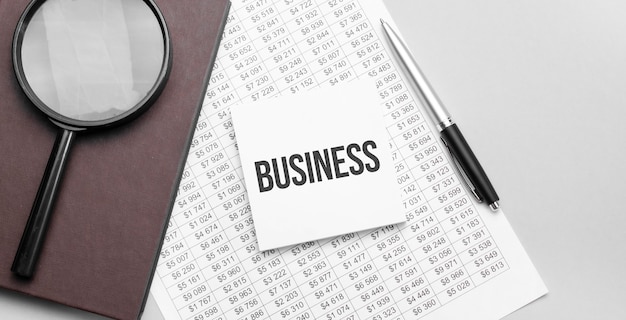 Magnifying glass, financial document, white paper sheet witn\
business sign and brown notebook on grey background.