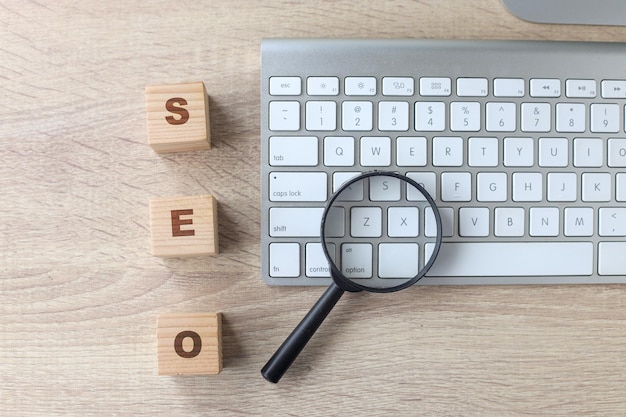 Magnifying glass on keyboard with wooden blocks lettering SEO on a wooden background.