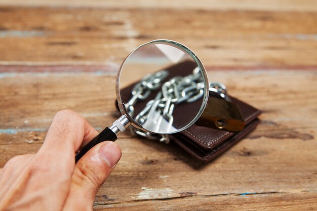 Magnifying glass looks at a wallet in chains, closed with a padlock on the table