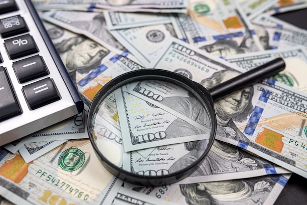 나무 테이블 세금 및 환급 검색 또는 재무 보고서 검토 개념에 격리된 계산기가 있는 미국 달러 지폐 더미에 돋보기
