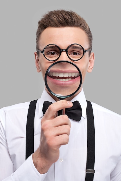 Magnifying smile. Portrait of cheerful young man in bow tie and suspenders holding magnifying glass in front of his mouth and smiling