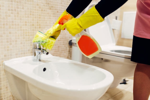 Le mani della cameriera in guanti di gomma puliscono il bidet con uno spray detergente, l'interno del bagno dell'hotel. servizio di pulizia professionale, donna di servizio, lavorazioni sanitarie