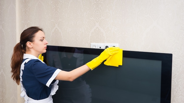 Photo maid or housekeeper cleaning a television set dusting the top with a yellow duster dressed in a neat uniform, profile upper body view