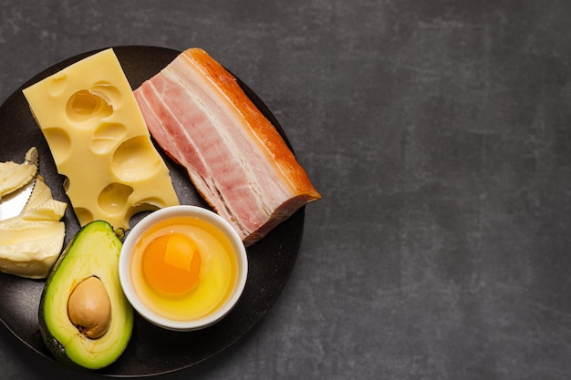 Main keto foods as butter olive oil fried egg avocado cheese fat meat bacon for ketogenic diet on black plate on dark rustic stone background products for healthy weight loss top view