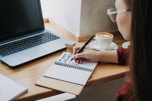 Photo main life goals how to set and achieve life goals young woman girl writing in notebook the inscription my life goals while sitting with laptop and papers in cafe