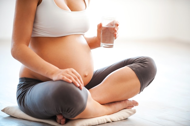 水のバランスを維持します。蓮華座に座っている間、水のガラスを保持している妊娠中の若い女性のクローズアップ