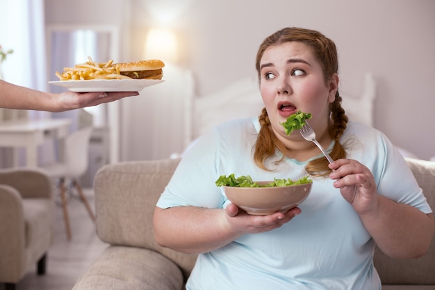 선택하십시오. 건강한 음식과 정크 푸드 사이에서 선택하는 동안 소파에 앉아 통통한 젊은 여성