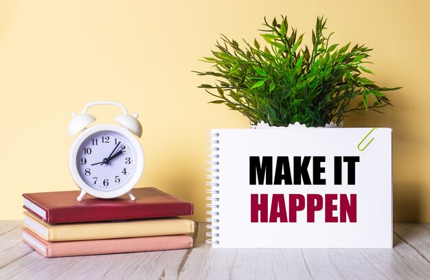 MAKE IT HAPPEN is written in a notebook next to a green plant and a white alarm clock, which stands on colorful diaries.