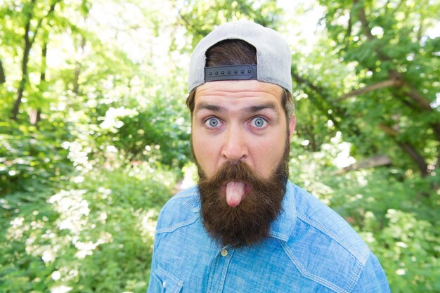 Make life worth living. summer day fun. bearded man looking funny. brunette bearded man wear cap in forest on background of trees. brutal brunette man making selfie. mature guy with beard in wood.