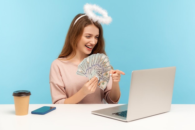 Photo make money on inteet angelic rich woman with nimbus holding dollar banknotes and pointing laptop screen while talking on video call encouraging to ea online indoor studio shot isolated