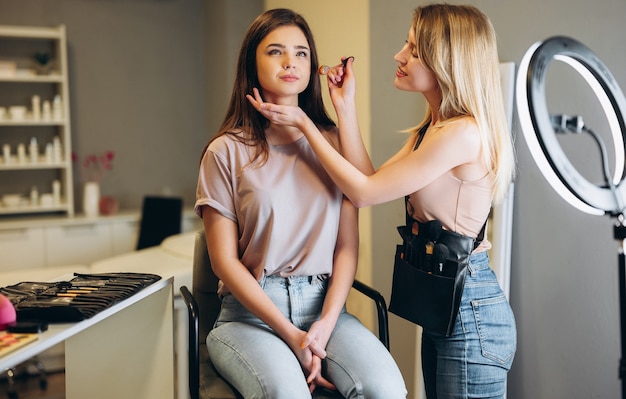 A make-up artist is applying makeup to a woman client. Woman with blond hair doing makeup to a woman with brown hair.