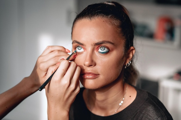 Make-up artist is bringing eyes with a blue shadow to a young beautiful lady with big grey amazing eyes, close up