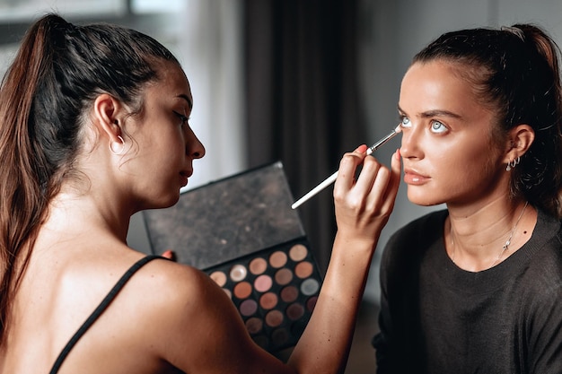 Make-up artist is doing make up to a young beautiful lady with big eyes, using palette of shadows