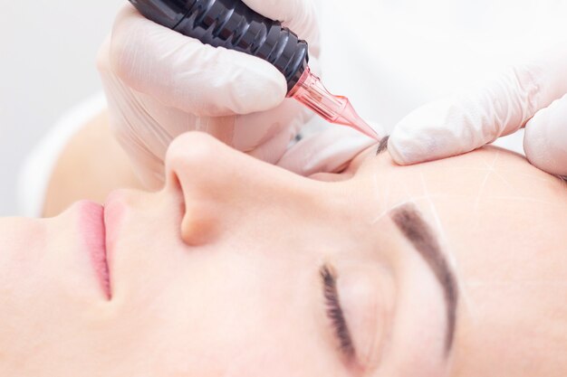 Make-Up. Beautician Hands Doing Eyebrow Tattoo On Woman Face.Permanent Brow Makeup In Beauty Salon. Closeup Of Specialist Doing Eyebrow Tattooing For Female. Cosmetology Treatment.