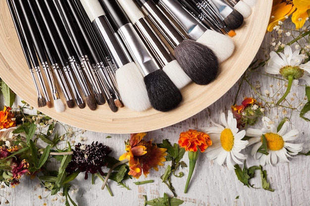 Make up brushes on a plate next to wild flowers on wooden background