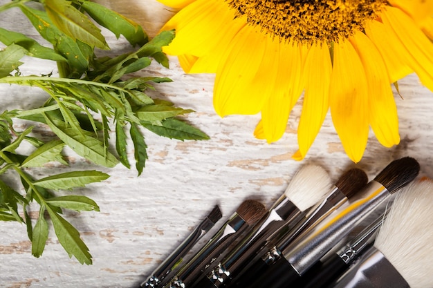 Make up brushes on a table next to wild flowers on wooden background