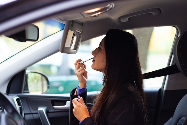 Make-up doen in de auto, zijaanzicht van een mooie zakenvrouw die achter het stuur zit en lippenstift gebruikt