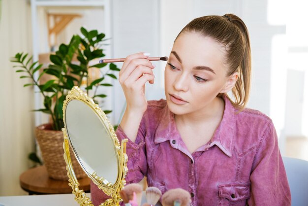 Make-up thuis jonge mooie vrouw met make-up borstel past oogschaduw toe voor een kleine spiegel...