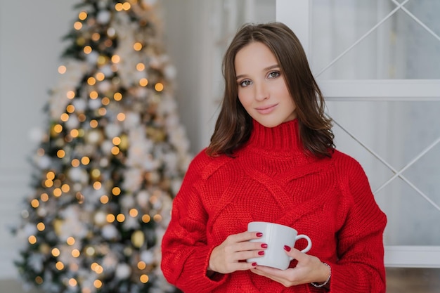 Make-up vrouw in gezellige winter trui houdt koffie plannen vakantie door Nieuwjaarsboom rust concept
