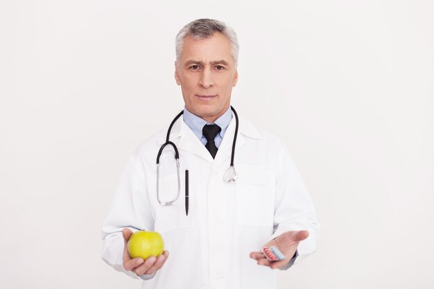 Fate la vostra scelta! medico anziano dei capelli grigi in uniforme che tiene una mela in una mano e delle pillole in un'altra mentre sta in piedi isolato su bianco