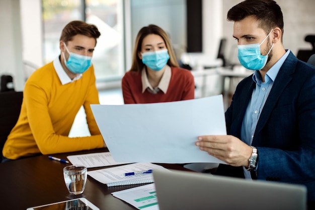 Makelaar in onroerend goed en een stel analyseren huisvestingsplannen tijdens een vergadering terwijl ze beschermende gezichtsmaskers dragen