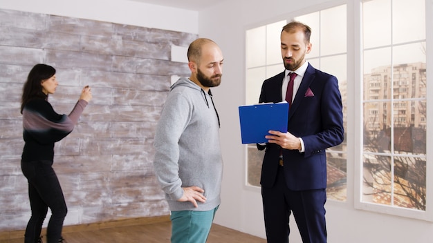 Makelaar in pak beschrijft appartement aan klant in leeg pand terwijl vrouw foto's maakt op de achtergrond.