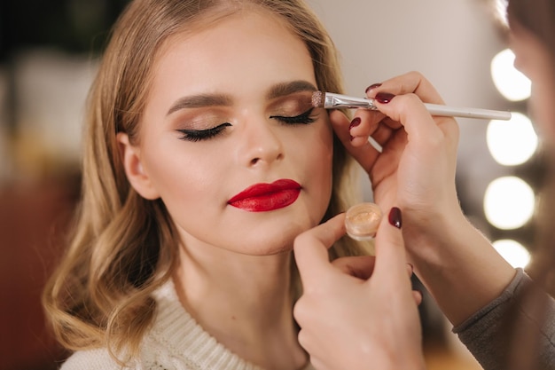 Makeup artist applies eye shadow perfect evening makeup Beauty blond hair girl with perfect skin and red lipstick