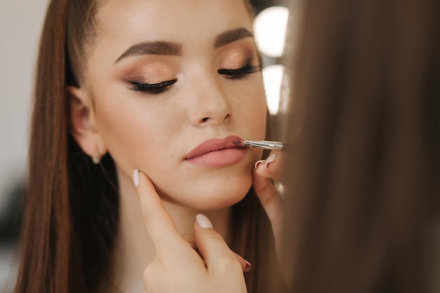 Makeup artist applies lipstick hand of makeup master painting lips of young beauty redhead model