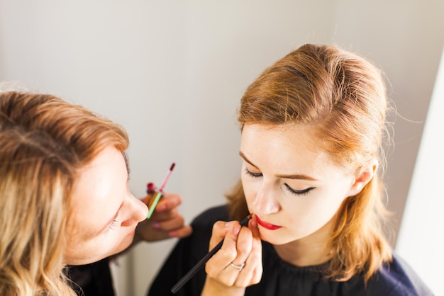 메이크업 아티스트는 빨간 립스틱을 바릅니다. 메이크업 마스터의 손, 젊은 뷰티 모델 소녀의 입술을 페인팅합니다.